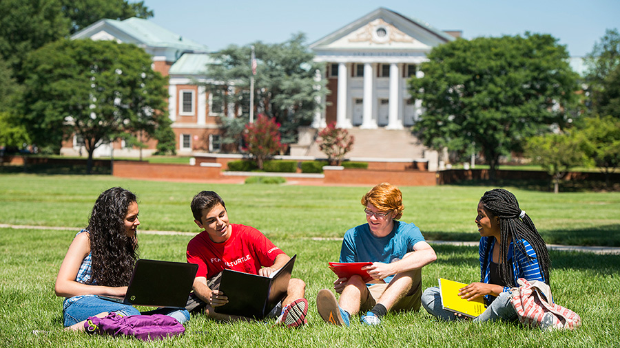four students sitting in the grass chatting in front of a campus building