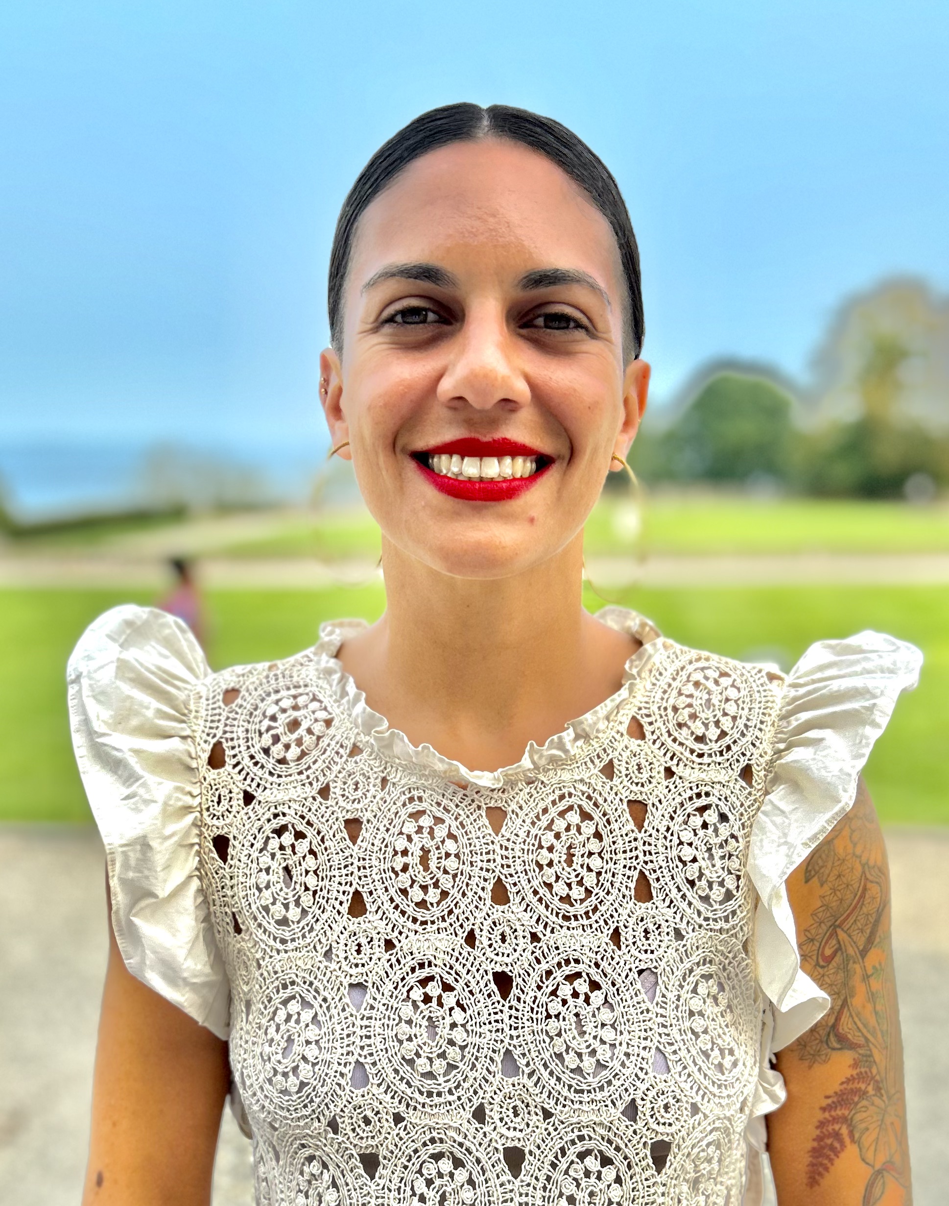 Dr. Zocal Da Silva is outdoors on a sunny day, with a blue sky. She has light brown skin, hazel eyes, red lipstick, and her brown hair is tied. She wears a beige blouse.